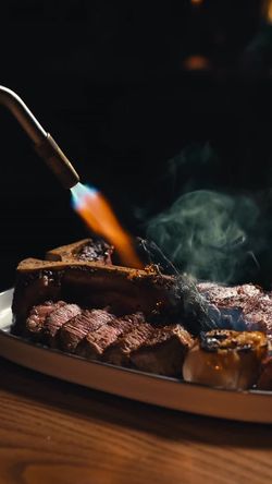 Flame searing a thick cut steak on a plate with grilled garlic on a wooden table at a fine dining restaurant.