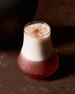Cocktail glass with a layered drink, featuring a reddish bottom and a thick foamy white top, sprinkled with spices.