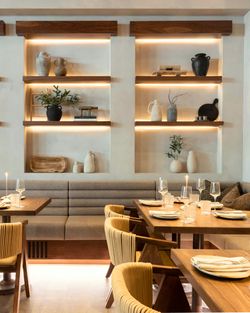 Mediterranean restaurant interior with wooden tables, beige cushioned seating, decorative shelves, and warm lighting.