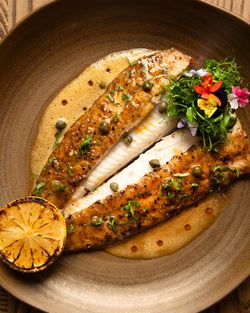 Grilled fish fillets garnished with capers and herbs, served with a grilled lemon half and a small salad with edible flowers.
