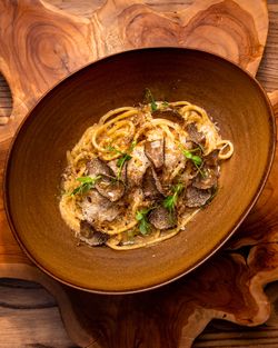 Creamy pasta with shaved black truffles and microgreens served in a brown bowl on a wooden board.