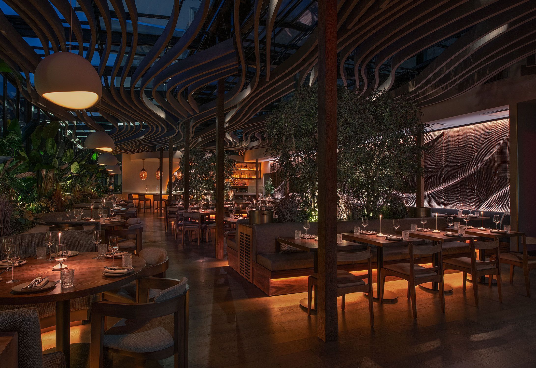 Fine dining restaurant interior with wooden tables, ambient lighting, decorative ceiling, and greenery at night
