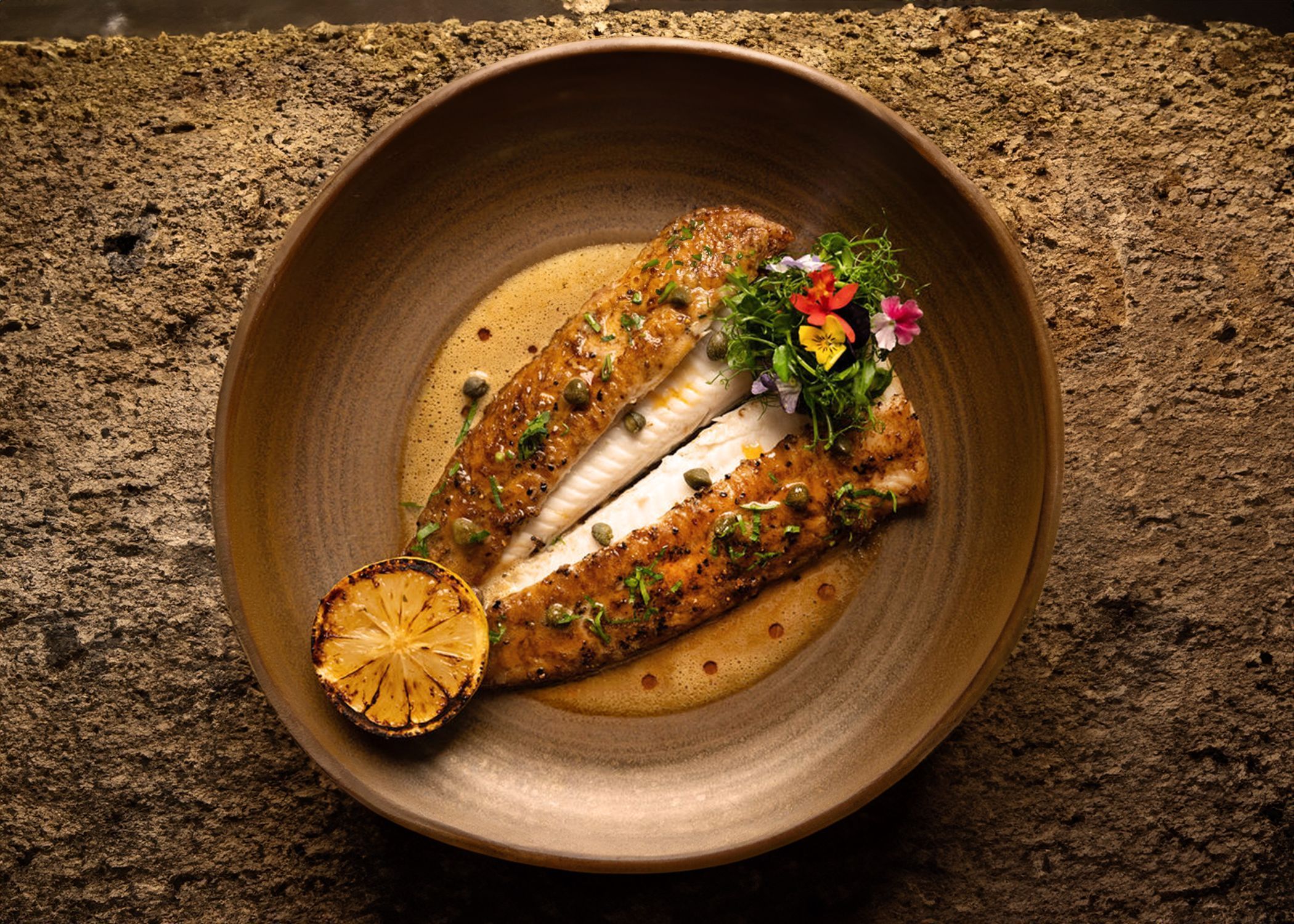 Grilled fish fillet with capers, lemon half, and herbs served on a brown ceramic plate at Mediterranean fine dining.