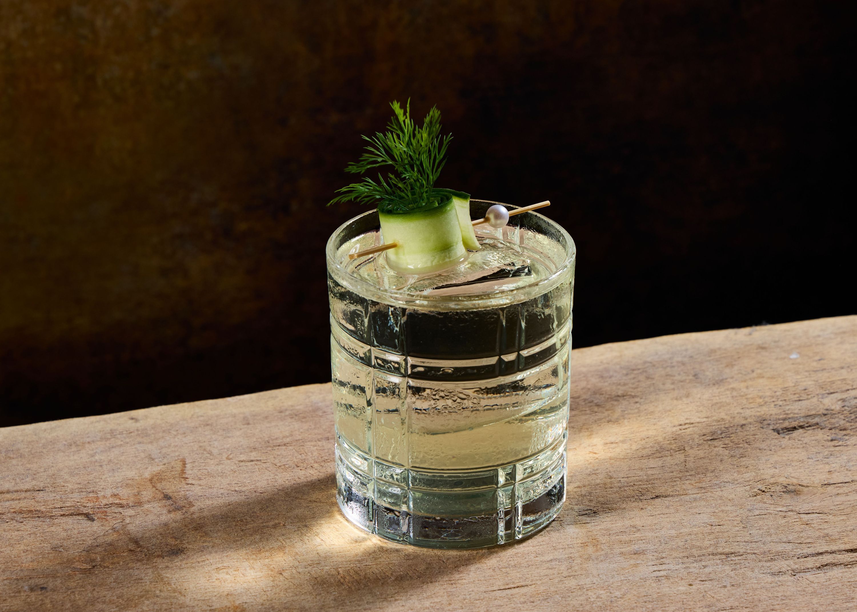 Clear cocktail garnished with cucumber ribbon and fresh herb served in a cut-glass tumbler on wooden surface