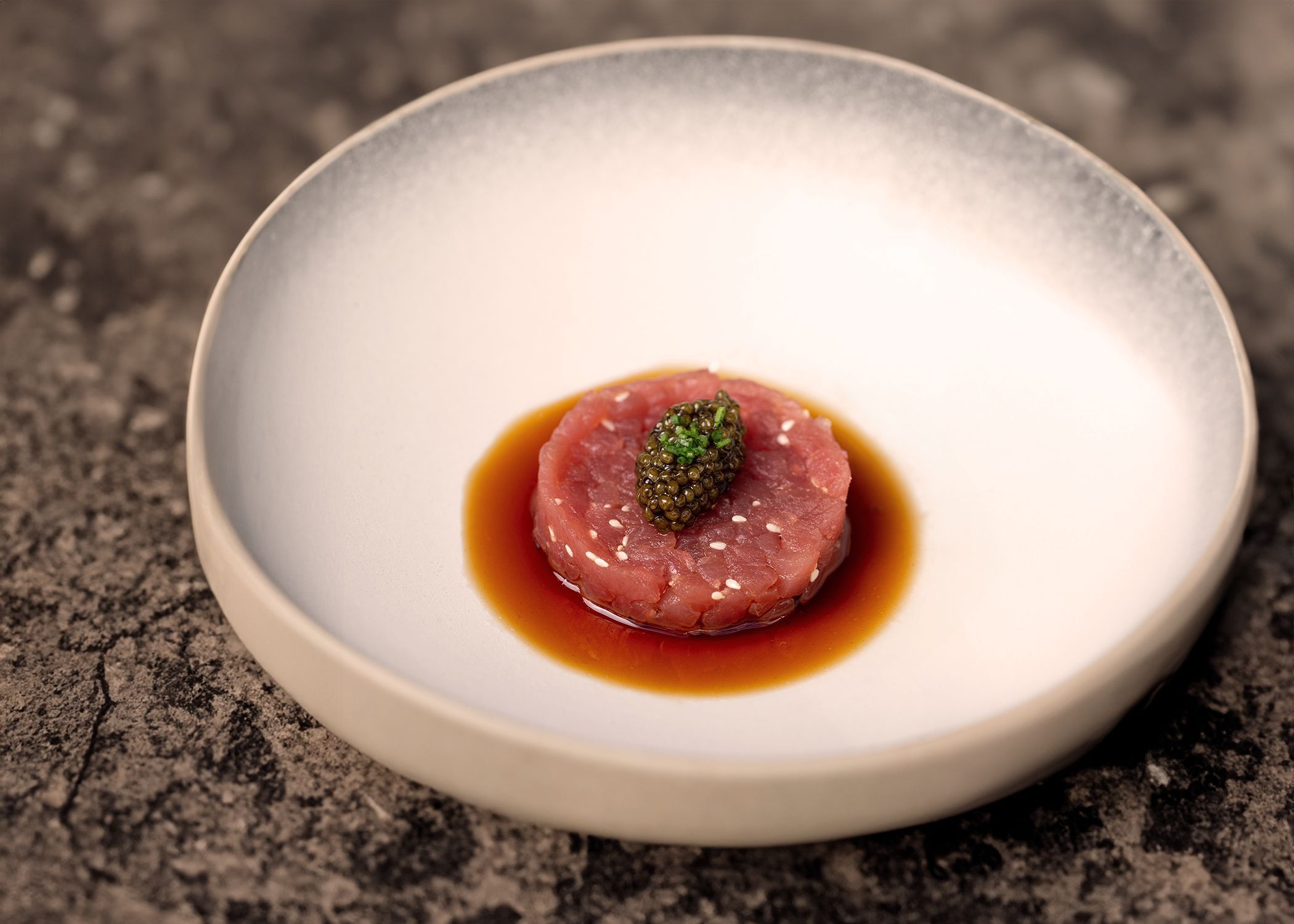 A fine dining dish featuring a round serving of raw tuna topped with caviar and chives, presented in a shallow sauce bowl.