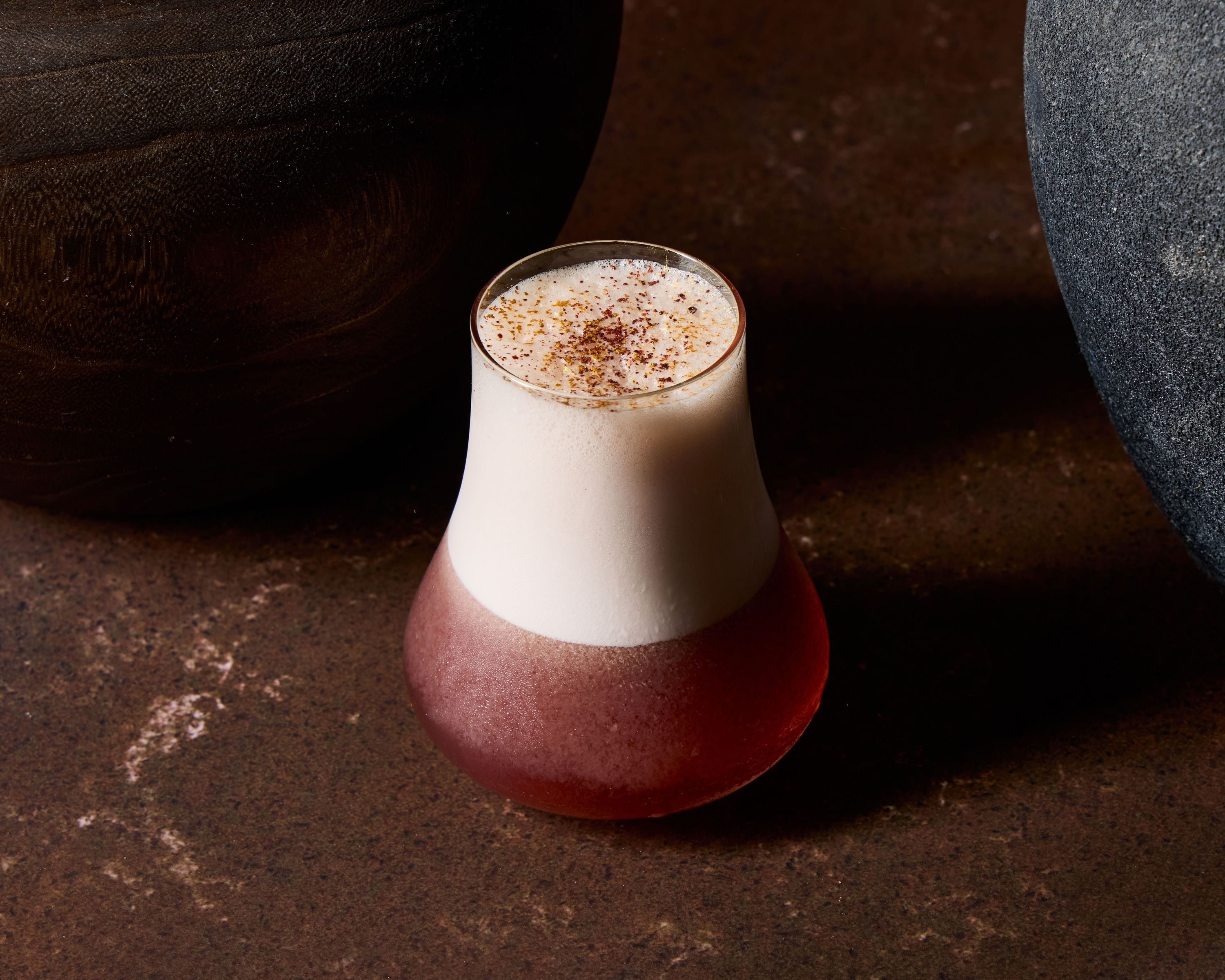 Cocktail with frothy top and sprinkled spices served in a curvy glass, placed on a rustic brown surface between stones.