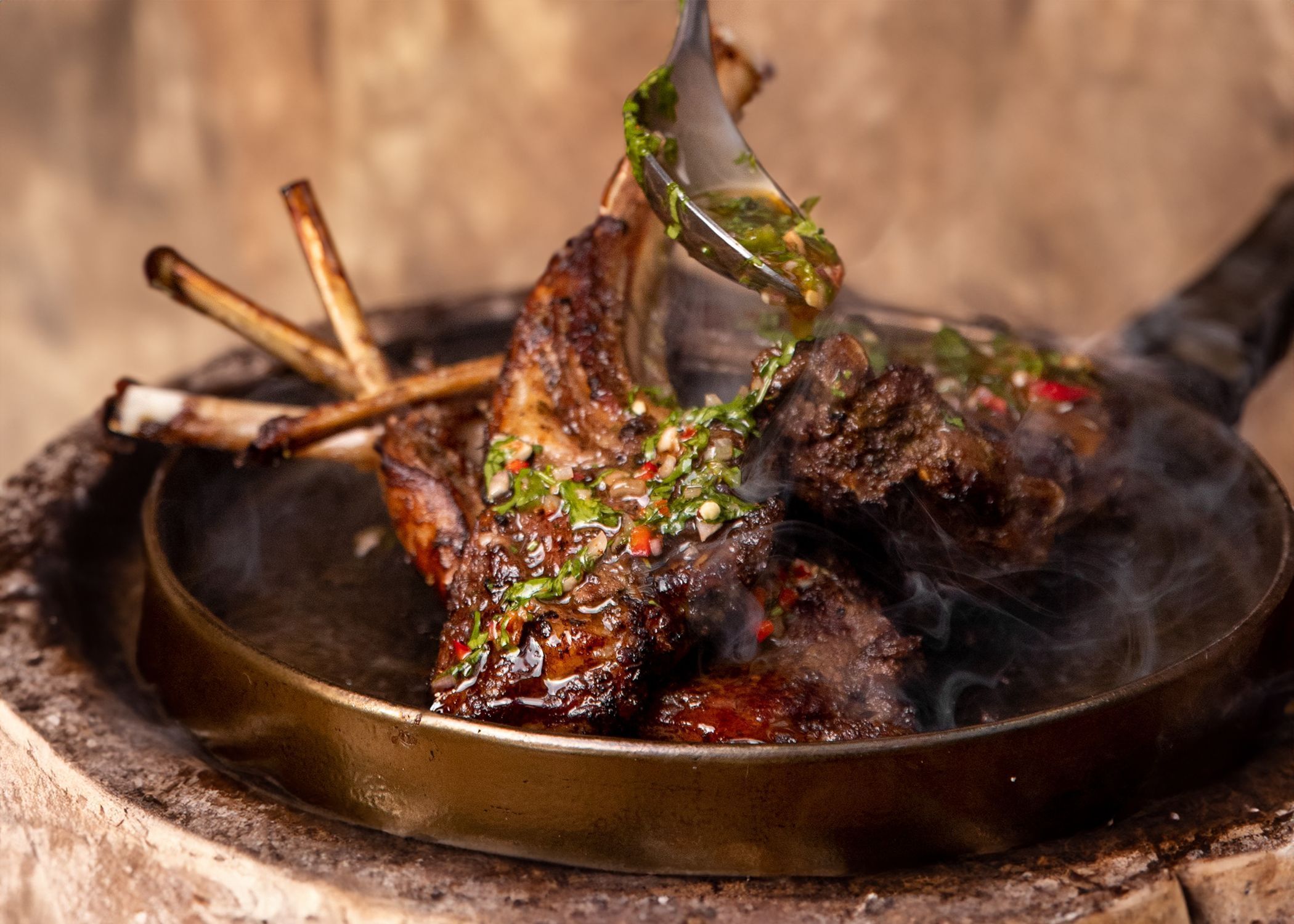 Grilled rack of lamb being garnished with a fresh herb sauce in a round pan at a fine dining Mediterranean restaurant.